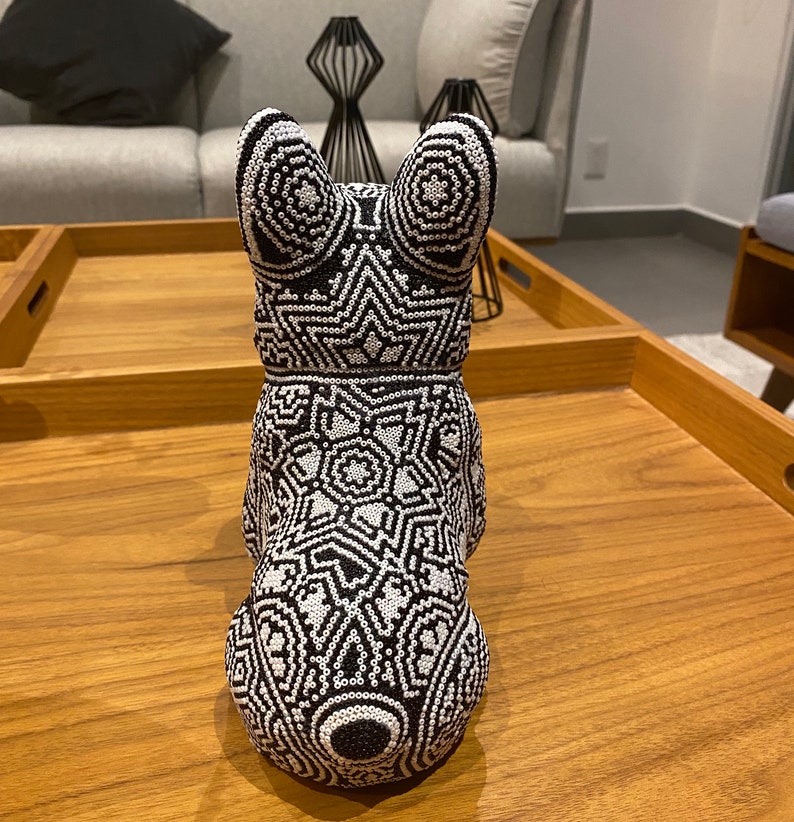 May include: A black and white beaded figurine of a dog. The figurine is sitting on a wooden surface. The dog is wearing a geometric pattern.
