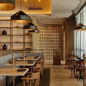 May include: A modern restaurant interior with wooden tables and chairs, a gray banquette, and a wooden slat wall.  The restaurant has large windows and black pendant lights.