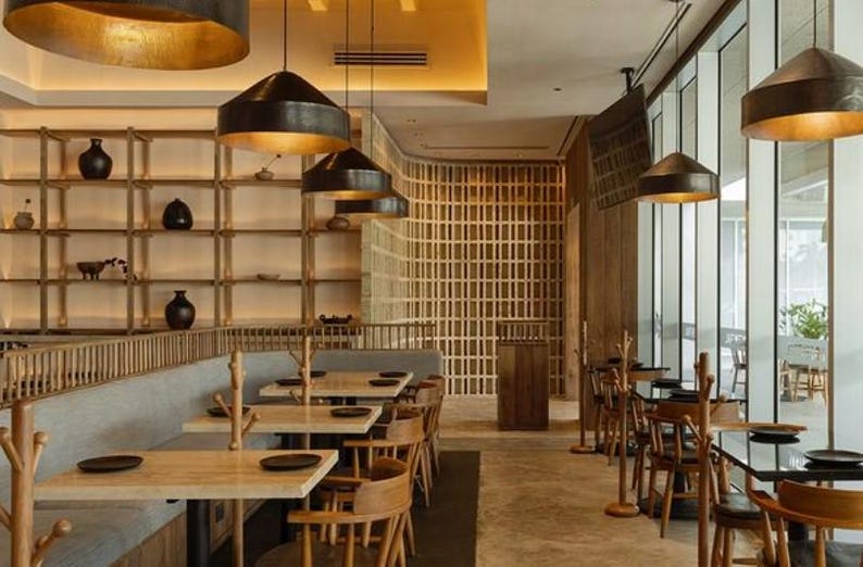 May include: A modern restaurant interior with wooden tables and chairs, a gray banquette, and a wooden slat wall.  The restaurant has large windows and black pendant lights.