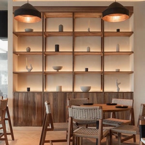 May include: A wooden shelving unit with a light wood finish and a white background. The shelves are filled with various decorative items, including vases, bowls, and sculptures. The unit is illuminated by two black pendant lights with gold accents. The shelves are in a modern style and would be a great addition to any home.