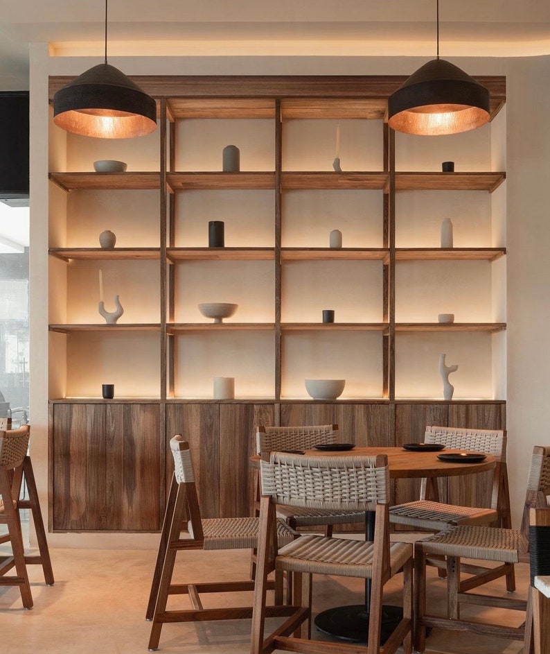 May include: A wooden shelving unit with a light wood finish and a white background. The shelves are filled with various decorative items, including vases, bowls, and sculptures. The unit is illuminated by two black pendant lights with gold accents. The shelves are in a modern style and would be a great addition to any home.