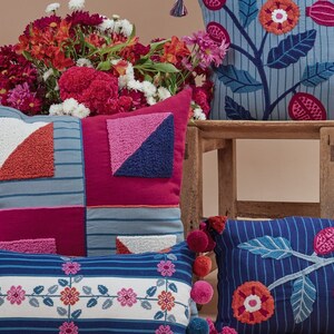 May include: A set of four decorative throw pillows with embroidered floral and geometric patterns. The pillows are in shades of blue, pink, red, and white. The pillows are arranged on a wooden table with a bouquet of flowers in the background.