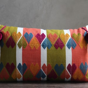 May include: A rectangular pillow with a colorful geometric pattern of hearts and stripes in shades of green, red, white, blue, yellow, and pink. The pillow has tassels on each end with pom-poms.