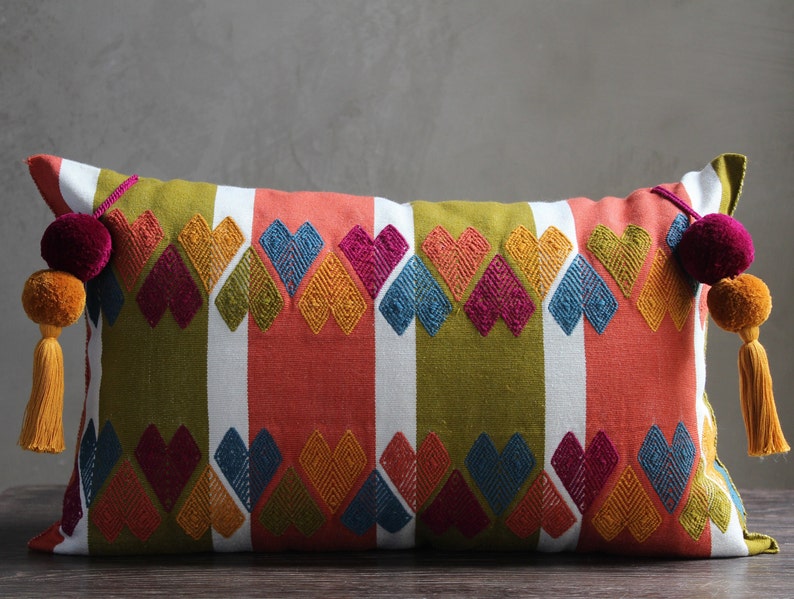 May include: A rectangular pillow with a colorful geometric pattern of hearts and stripes in shades of green, red, white, blue, yellow, and pink. The pillow has tassels on each end with pom-poms.