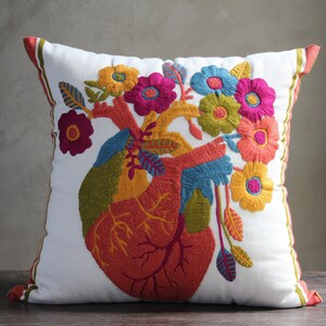 May include: White pillow with a colorful embroidered heart design. The heart is surrounded by flowers in shades of pink, yellow, orange, and blue. The pillow has a white background and a brown border.
