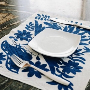 May include: A white ceramic plate with a square shape sits on a white placemat with a blue floral pattern. The placemat has a blue embroidered design of flowers and leaves.