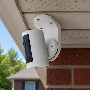 May include: A white security camera with a black front panel is mounted on a white bracket. The camera is attached to a red brick wall and a beige soffit. The sky and green trees are visible in the background.