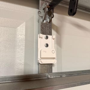 May include: A white plastic mounting bracket for a Ring device, secured with two silver screws to a vertical metal garage door track. The bracket features a rectangular base with a small circular cutout and the word ring printed in gray.