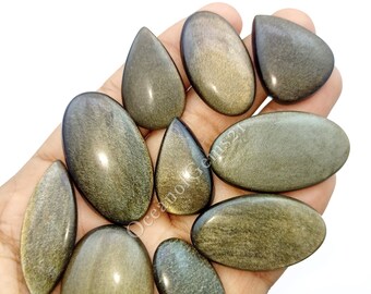 Könnte beinhalten: Eine Sammlung von zehn Goldschimmer-Obsidian-Cabochons in verschiedenen Formen und Größen. Die Cabochons sind eine Mischung aus ovalen und tropfenförmigen Formen. Die Steine haben einen goldenen Schimmer und eine dunkelbraune Basis.
