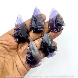 Può includere: Cinque punte di freccia di ametista, ciascuna con una forma e una dimensione diverse, sono mostrate su una mano. Le punte di freccia sono di colore viola scuro con un leggero bagliore iridescente.