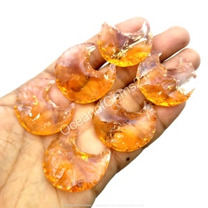May include: Six orange and clear glass cabochons in the shape of a crescent moon. The cabochons are in a hand, with the text "OceanofGems21" visible.