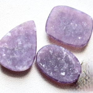 May include: Three purple lepidolite gemstone cabochons in various shapes, including a teardrop, oval, and rectangular shape. The stones have a light purple color with a sparkly, iridescent finish.