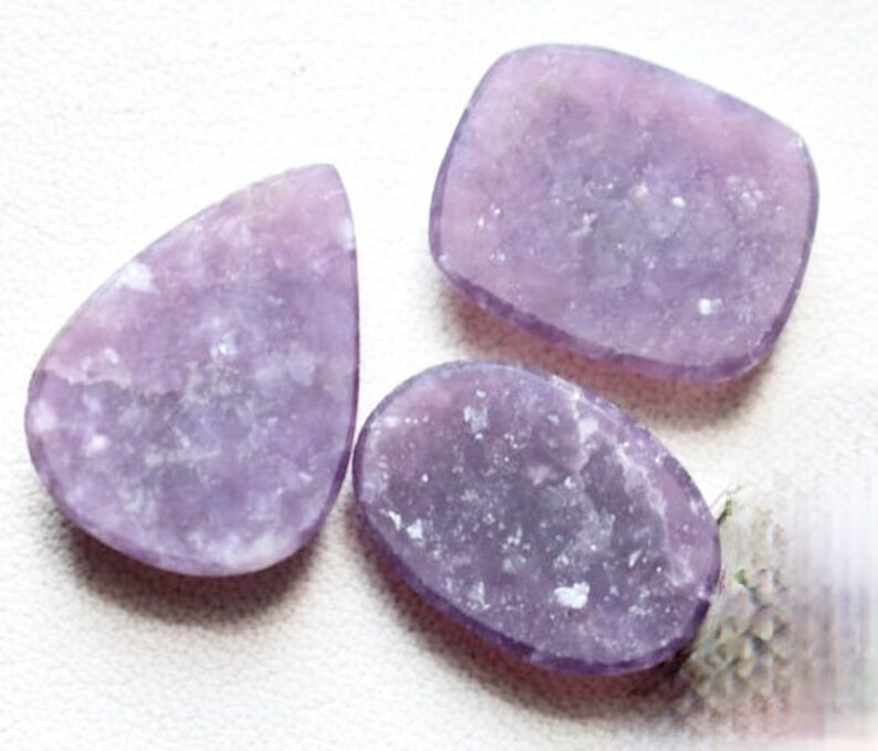 May include: Three purple lepidolite gemstone cabochons in various shapes, including a teardrop, oval, and rectangular shape. The stones have a light purple color with a sparkly, iridescent finish.