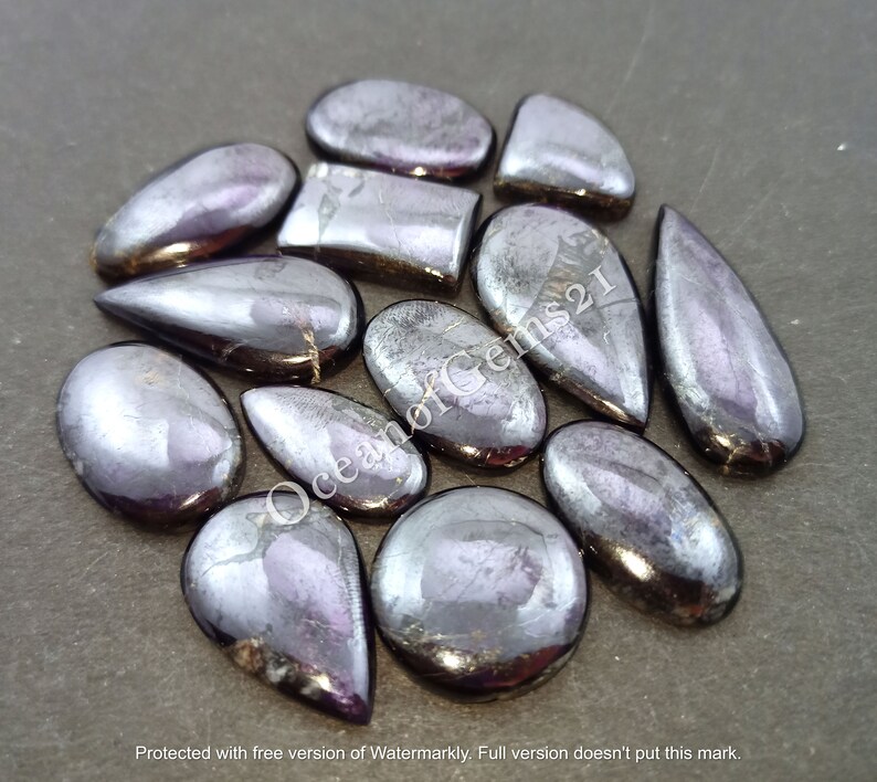 K&ouml;nnte beinhalten: Eine Sammlung von 12 regenbogenfarbenen schwarzen Edelstein-Cabochons in verschiedenen Formen, darunter Tropfen, Oval und Rechteck. Die Steine haben eine schimmernde, metallische Oberfl&auml;che.