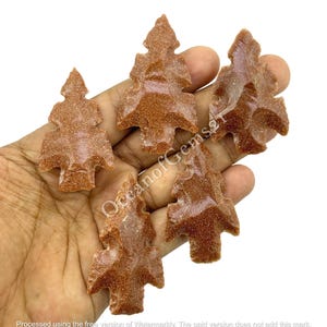 May include: Four brown sandstone arrowhead shaped crystals in the form of a Christmas tree. The crystals are in a hand, with the tips of the trees pointing upwards.