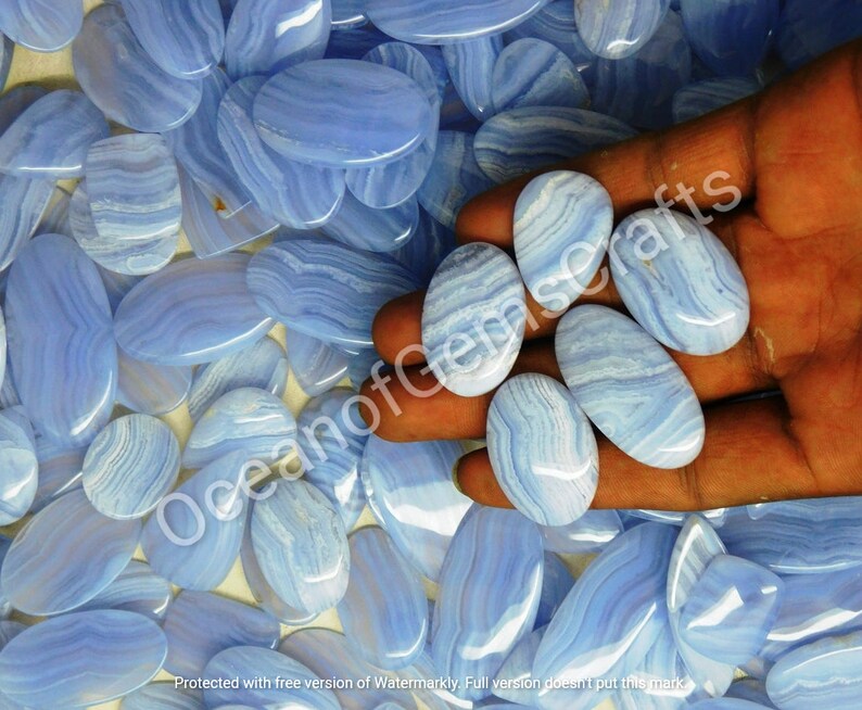 May include: A close-up of a hand holding several oval-shaped blue agate stones. The stones have a light blue and white banded pattern. The stones are surrounded by more of the same stones.