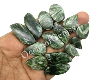 Puede incluir: Una colección de piedras preciosas de serafinita verde en varias formas, incluyendo ovaladas, de lágrima e irregulares. Las piedras muestran un patrón de plumas distintivo y se sostienen en una mano. El texto "Oceanof Gems21" es visible.