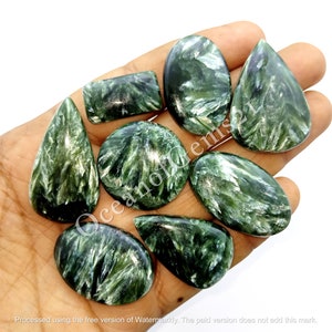 May include: A collection of polished green seraphinite gemstones in various shapes, including oval, teardrop, and rectangular. The stones display a feathery, silvery pattern, held in a hand against a white background. The gemstones are of different sizes.