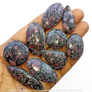 Peut inclure: Une collection de neuf cabochons ovales et en forme de goutte de pierre mouchetée de noir et de rose. Les pierres sont lisses et polies.