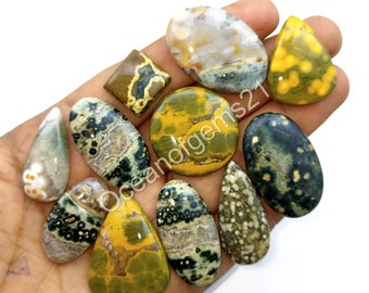 May include: Assortment of polished ocean jasper cabochons in various shapes and sizes. The cabochons feature a mix of green, yellow, brown, and white colors with black and white spots.