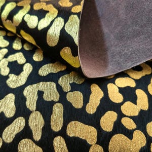 Può includere: Un pezzo di pelle animale nera con un motivo leopardato color oro metallizzato. Il materiale presenta una superficie testurizzata e pelosa sul lato anteriore e una finitura liscia in camoscio marrone opaco sul retro dove la pelle è ripiegata.