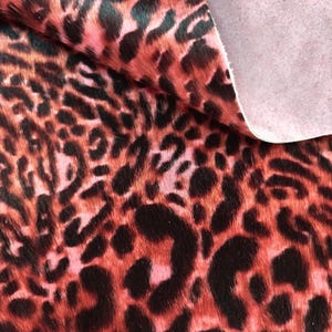 Könnte beinhalten: Ein Stück Tierhautleder mit einem leuchtend roten und schwarzen Leopardenmuster. Die Oberfläche hat ein kurzes, strukturiertes, fellartiges Finish, während die Unterseite an der freiliegenden Ecke eine glatte, helle, matte Wildledertextur zeigt.
