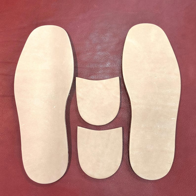 May include: A collection of beige leather shoe insoles and heel pieces, arranged on a burgundy surface. The insoles are in the shape of feet, with two full-length insoles and two smaller heel pieces.