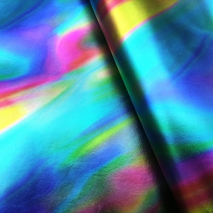 Pode incluir: Close-up de tecido iridescente com um espectro de cores que inclui azul, rosa, amarelo e verde. O material tem uma superfície lisa e refletora e está dobrado, criando sombras e destaques.