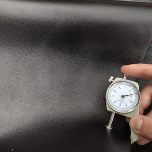 Op de afbeelding: Close-up van een zwart lederen materiaal met een meter die een dikte van 1,7 mm meet.
