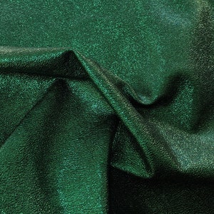 Puede incluir: Primer plano de una tela verde esmeralda doblada con un acabado texturizado y met&aacute;lico. La tela tiene un brillo sutil, y los pliegues crean sombras y reflejos, realzando la profundidad y la textura del material.