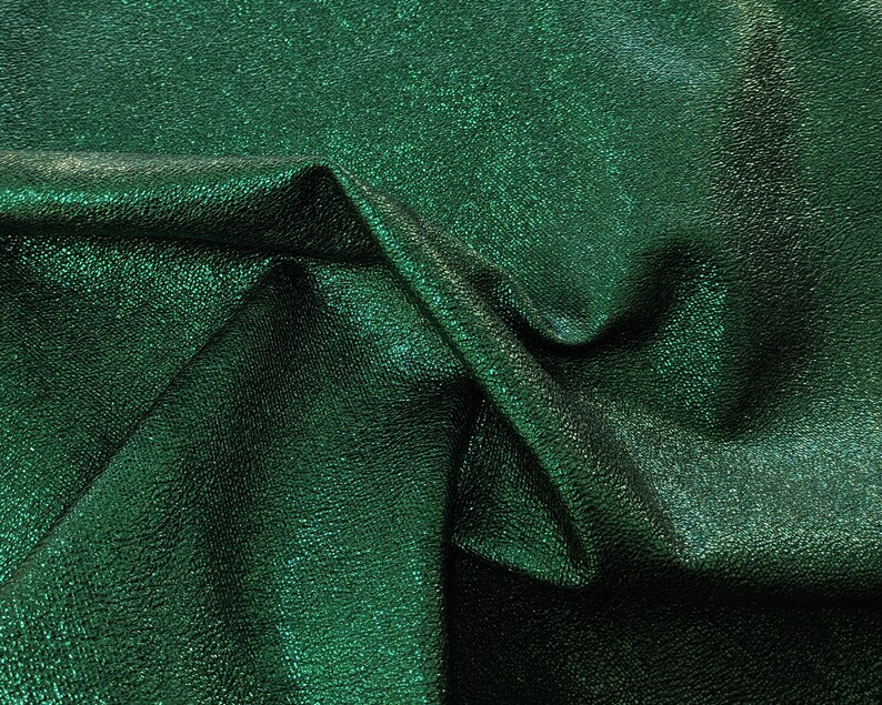 Puede incluir: Primer plano de una tela verde esmeralda doblada con un acabado texturizado y met&aacute;lico. La tela tiene un brillo sutil, y los pliegues crean sombras y reflejos, realzando la profundidad y la textura del material.