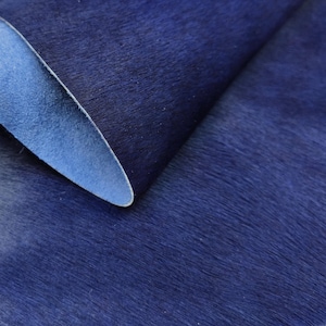 May include: Close-up of a sheet of deep blue dyed cowhide leather. The leather has a soft, furry texture and a smooth, light blue underside. The image highlights the rich color and natural grain of the material, suitable for crafting.