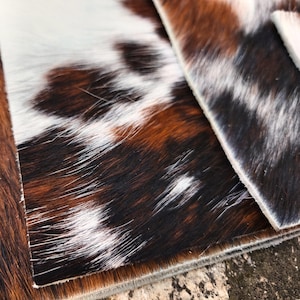 May include: Close-up of two pieces of cowhide leather. The leather has a natural pattern of brown, black, and white patches. The texture of the hair is visible, and the edges of the leather are neatly trimmed.