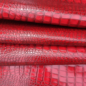 May include: Red crocodile embossed leather fabric. The fabric is folded in layers, showing the texture of the embossed pattern.