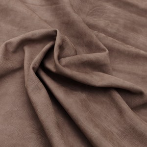 Pode incluir: Close-up de um peda&ccedil;o de couro castanho com uma superf&iacute;cie texturizada. O couro est&aacute; dobrado e drapeado, mostrando o gr&atilde;o natural e as varia&ccedil;&otilde;es de cor. O tom geral &eacute; um castanho suave e terroso.