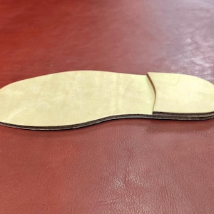 May include: A light-colored, wooden shoe sole with a separate heel piece. The sole is tan with a dark brown edge, and the heel is a lighter shade of tan. The sole is placed on a red surface.
