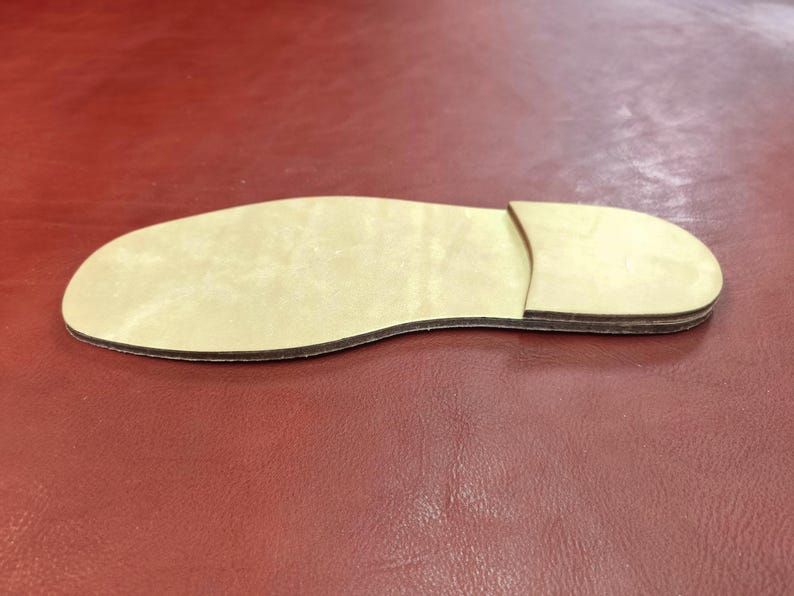 May include: A light-colored, wooden shoe sole with a separate heel piece. The sole is tan with a dark brown edge, and the heel is a lighter shade of tan. The sole is placed on a red surface.