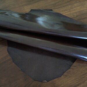 May include: A piece of dark brown leather with a glossy finish. The leather is folded in half and appears to be soft and supple.