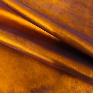May include: Close-up of a textured, metallic fabric with a gradient of colors. The fabric transitions from a deep, dark purple to a vibrant, golden orange. The material is folded, creating shadows and highlights that emphasize its texture.