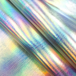 May include: Close-up of iridescent fabric with a textured surface. The material reflects a spectrum of colours, including blues, greens, and yellows, creating a shimmering effect. The fabric is folded, showing the layers and texture.