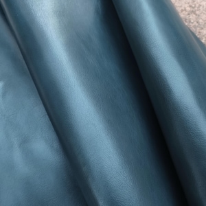 Pode incluir: Close-up de tecido de couro azul-petróleo com um brilho sutil. O material está dobrado, revelando a textura e a profundidade. O couro tem uma superfície lisa com ligeiras variações de cor.