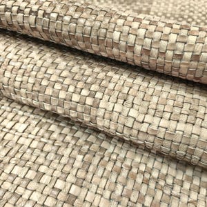 Puede incluir: Un material textil que presenta un patrón tejido con una estructura de rejilla. La superficie muestra una mezcla de tonos neutros, incluyendo matices de beige, marrón claro y blanco roto, creando un aspecto texturizado con tiras entrelazadas.