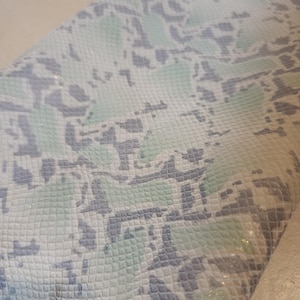 May include: Close-up of a textured material with a snakeskin pattern. The pattern features a mix of white, light blue, and grey tones. The material appears to be folded, revealing the texture and colour variations.