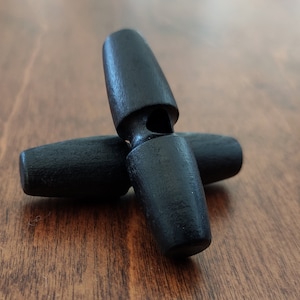 Puede incluir: Tres botones de madera negros con un agujero en el centro. Los botones están dispuestos en forma de cruz.