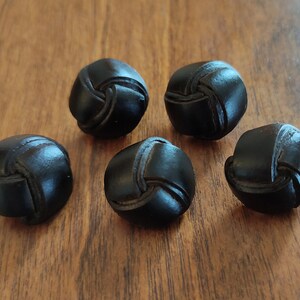 Peut inclure: Cinq boutons en cuir noir avec un design noué. Les boutons sont ronds et ont une finition lisse et polie.