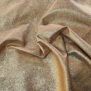 Pode incluir: Close-up de um tecido metálico dourado com superfície texturizada. O tecido está dobrado e drapeado, criando dobras e rugas suaves.