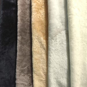 May include: Five vertical strips of plush faux fur fabric arranged side by side. The colours from left to right are deep black, muted taupe, warm beige, soft light grey, and creamy off-white. Each strip features a dense, soft, and fuzzy texture.