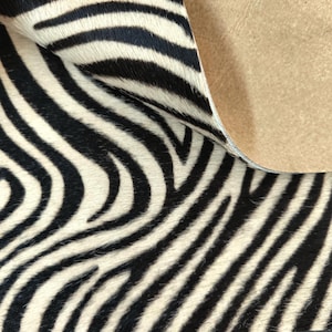 May include: A piece of animal hide featuring a black and white zebra stripe pattern. The material has a textured, hairy surface on the front side, while the reverse side shows a smooth, tan-colored suede backing visible where the corner is folded.