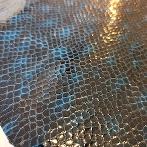 May include: Close-up of a textured material with a scale-like pattern. The material has a dark base colour with blue and gold highlights, creating a shimmering effect. The surface is uneven, with a slightly rough texture.