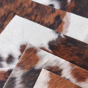 May include: Close-up of several pieces of cowhide leather. The leather has a natural, mottled pattern in shades of brown, white, and black. The texture appears soft and slightly raised, suggesting a high-quality material.